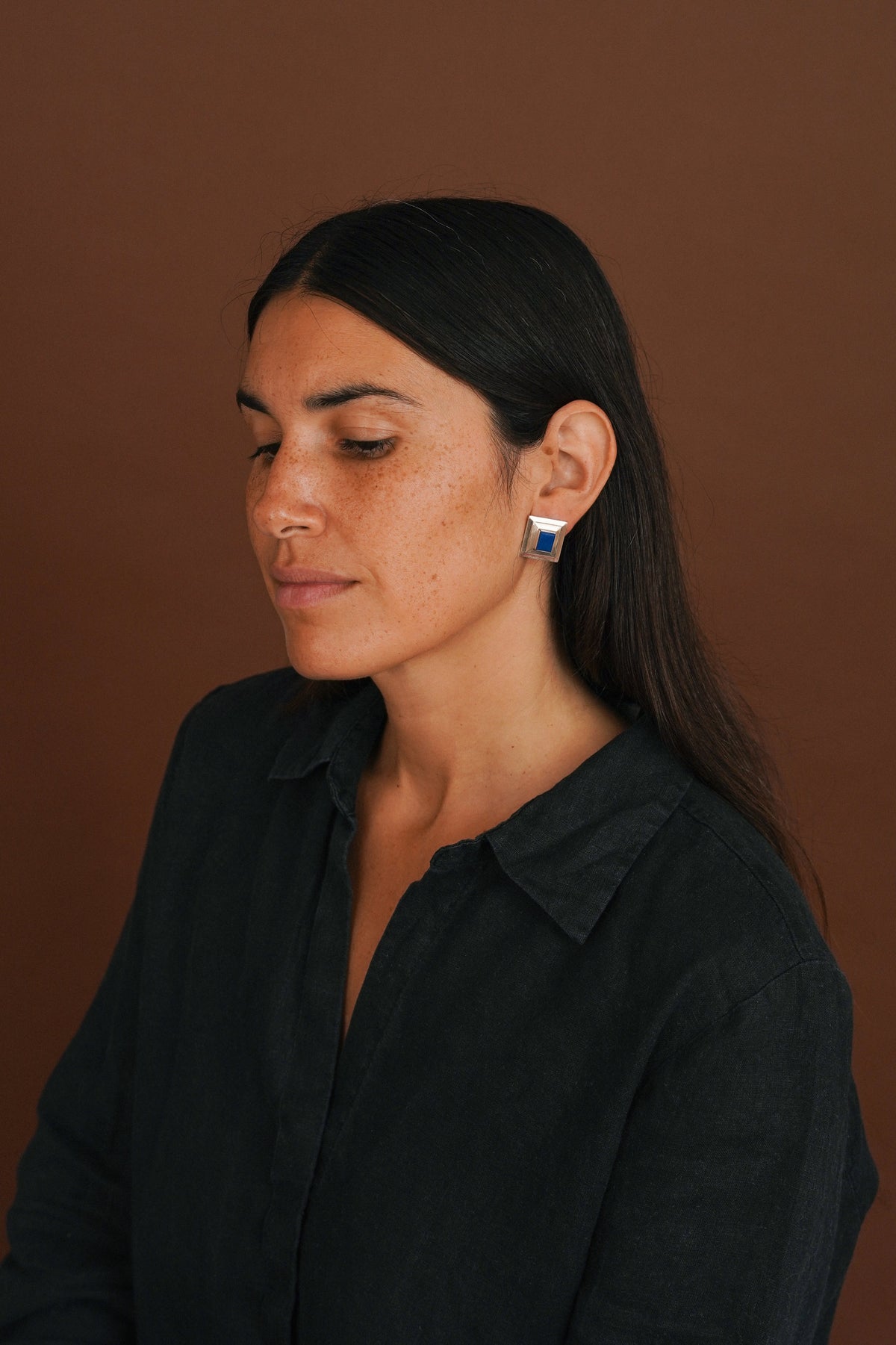 A woman with long dark hair is shown in profile against a solid warm brown background, wearing a small silver geometric stud earring with a blue and white inlay. She is wearing a dark black linen button-up shirt.