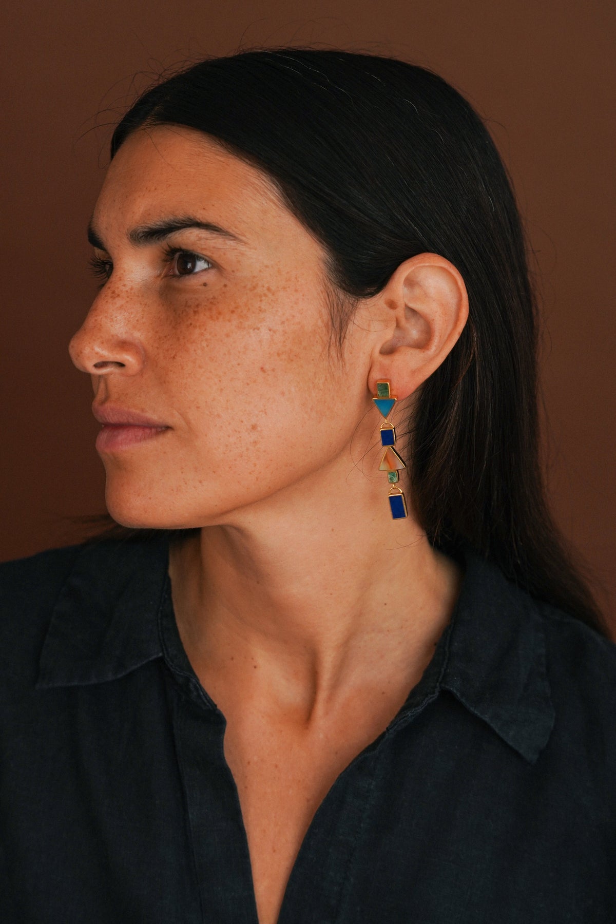 A close-up side profile of a woman with dark hair wearing a dark navy shirt and a pair of colorful geometric dangle earrings. The earrings feature blue, green, and gold elements against a solid warm brown background.