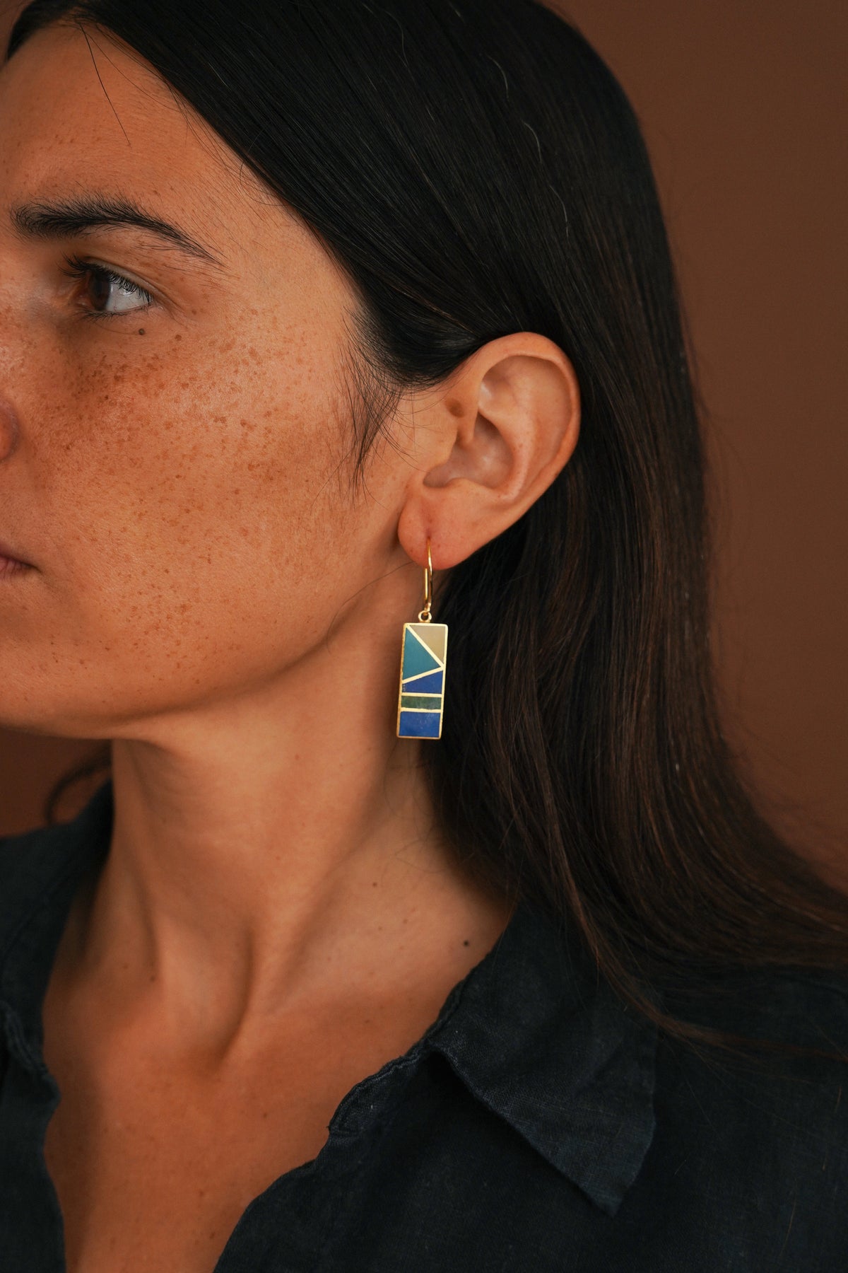 A close-up side profile of a woman with dark hair wearing a gold-tone dangle earring with a blue and yellow geometric patterned pendant, set against a warm brown background.