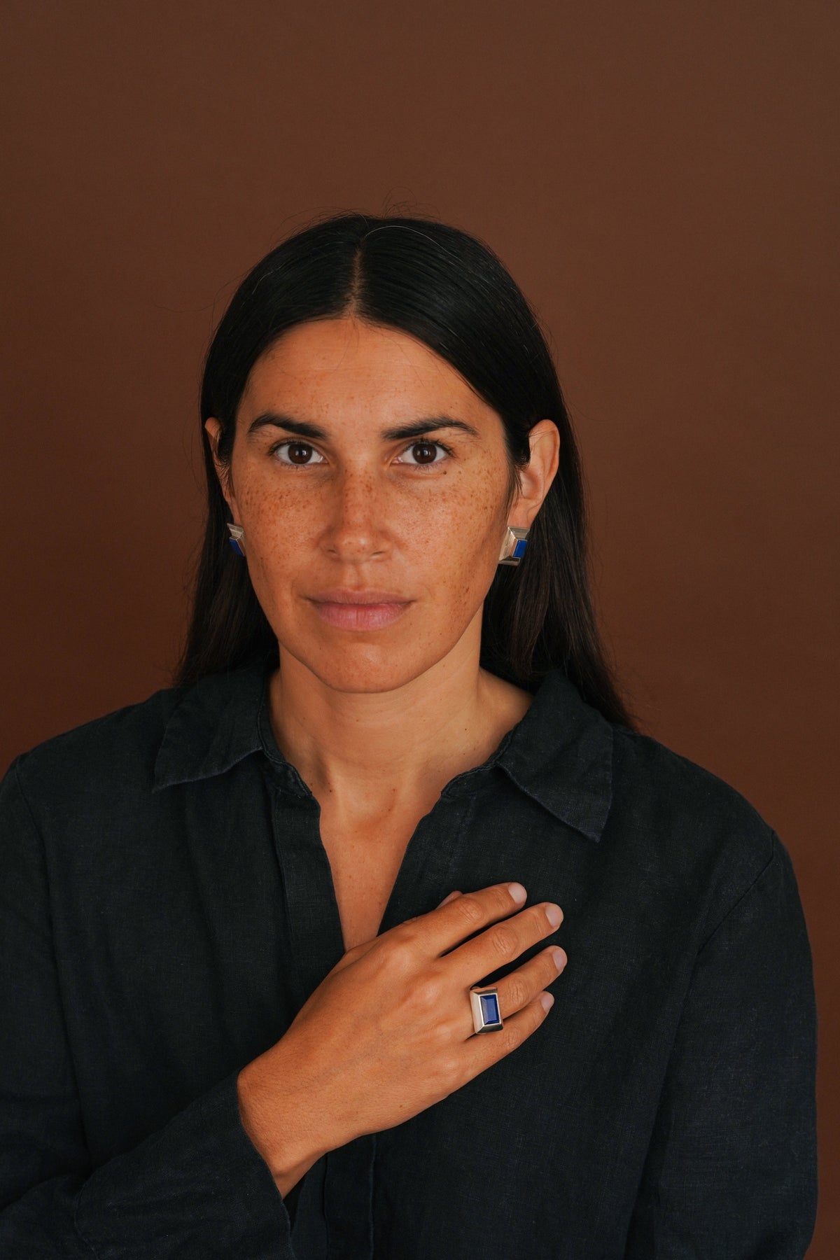 woman with dark hair wears a chunky lapis ring and a dark shirt, with a brown background