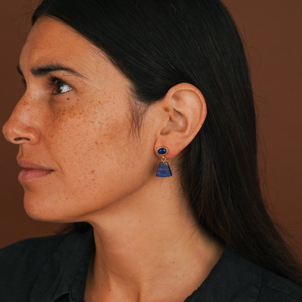 A close-up side profile of a woman with dark hair wearing a small gold hoop earring with a blue lapis lazuli dangling charm, set against a warm brown background.