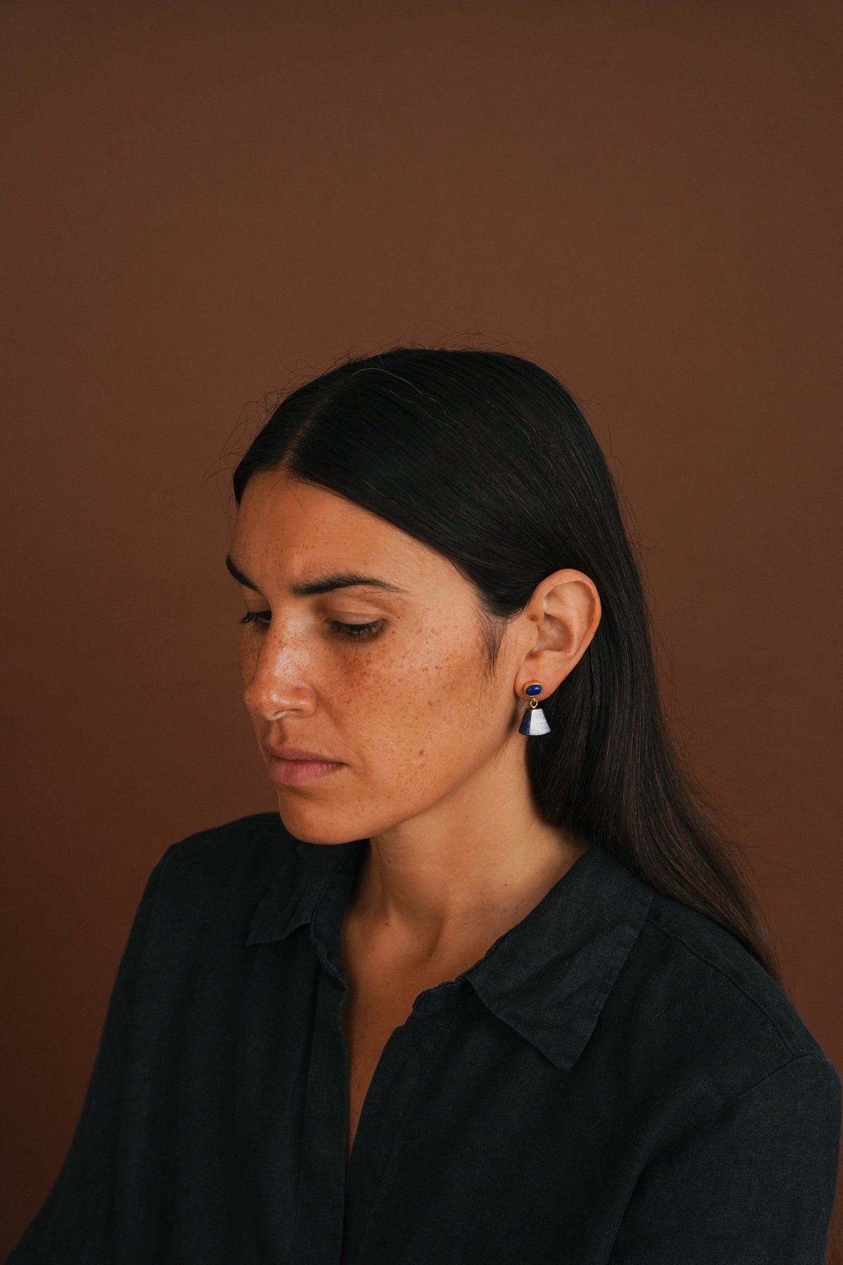 A woman with long dark hair wearing a black shirt and a small geometric dangle earring, shown in a close-up portrait against a solid brown background.