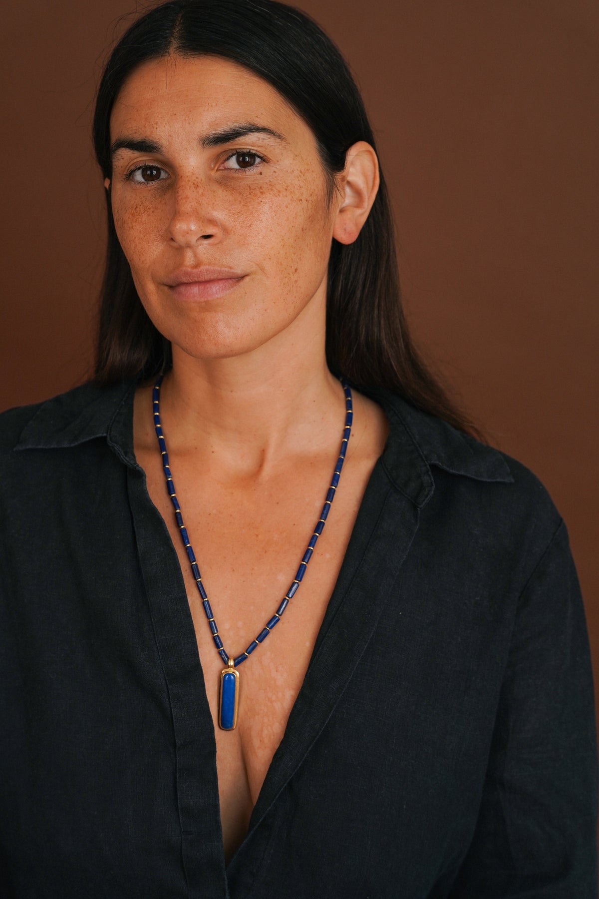 A woman with dark hair and freckles wearing a long beaded necklace with a blue rectangular pendant, photographed against a warm brown studio background.