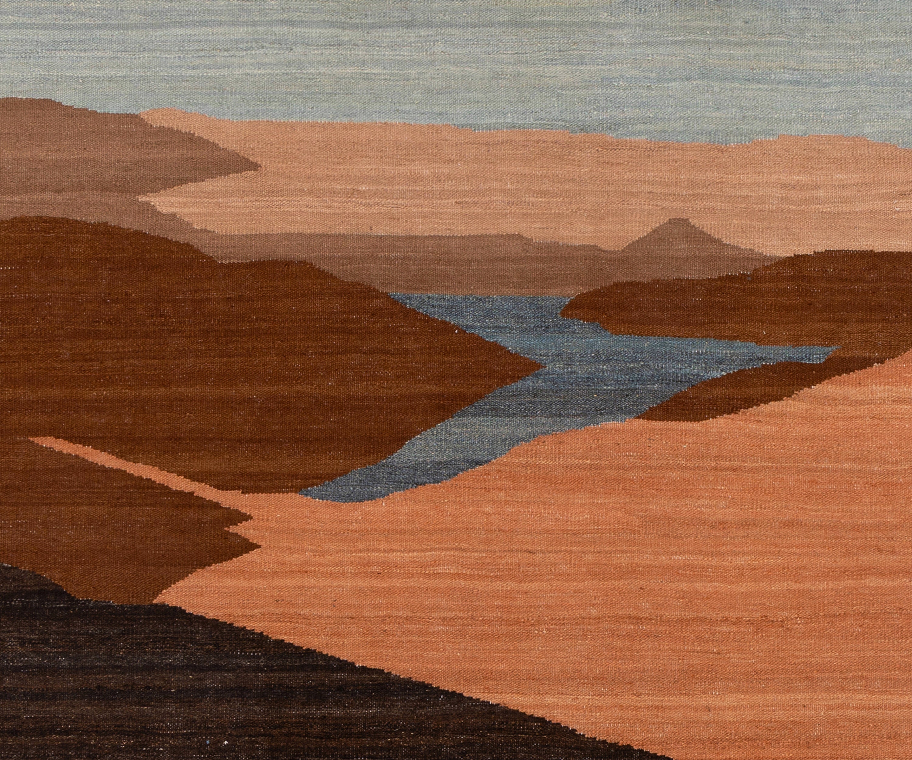 A close-up shot of a woven rug featuring a stylized landscape design with layered terracotta hills, a winding slate-blue river, and a muted gray-blue sky.