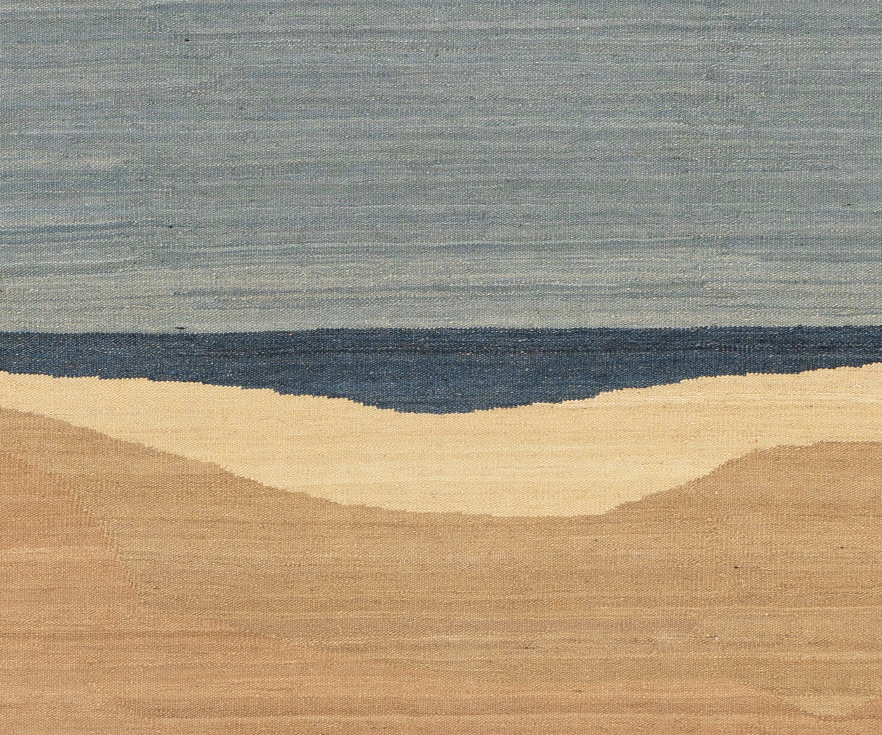 A close-up of a woven textile or rug pattern showing a horizontal landscape design with a light gray upper section, a dark blue middle band, and warm beige lower sections.