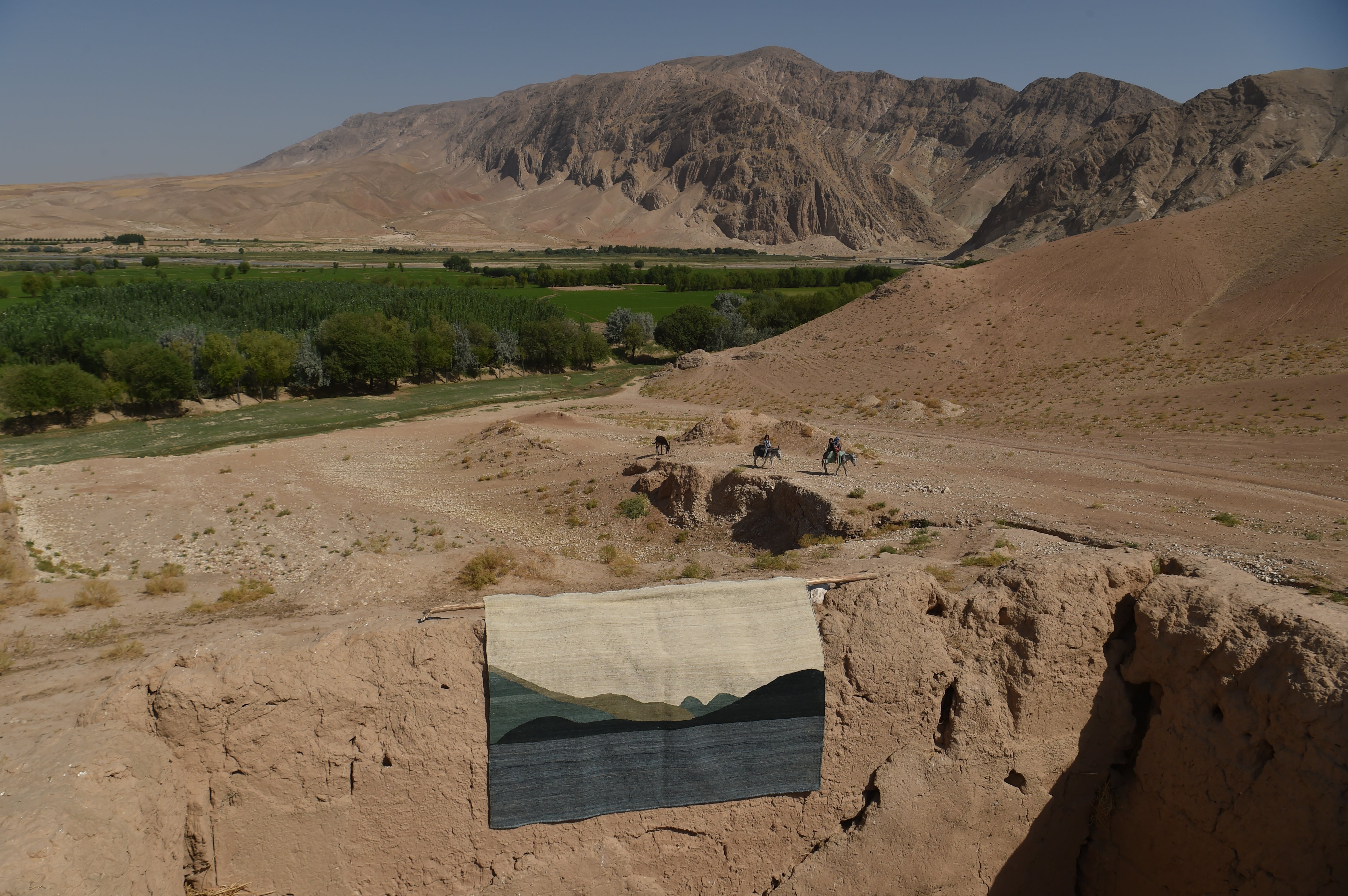 A landscape flatweave rug depicting a mountain and lake scene, displayed hanging on a sandy earthen wall in an outdoor desert setting with mountains and green fields in the background, Afghanistan.
