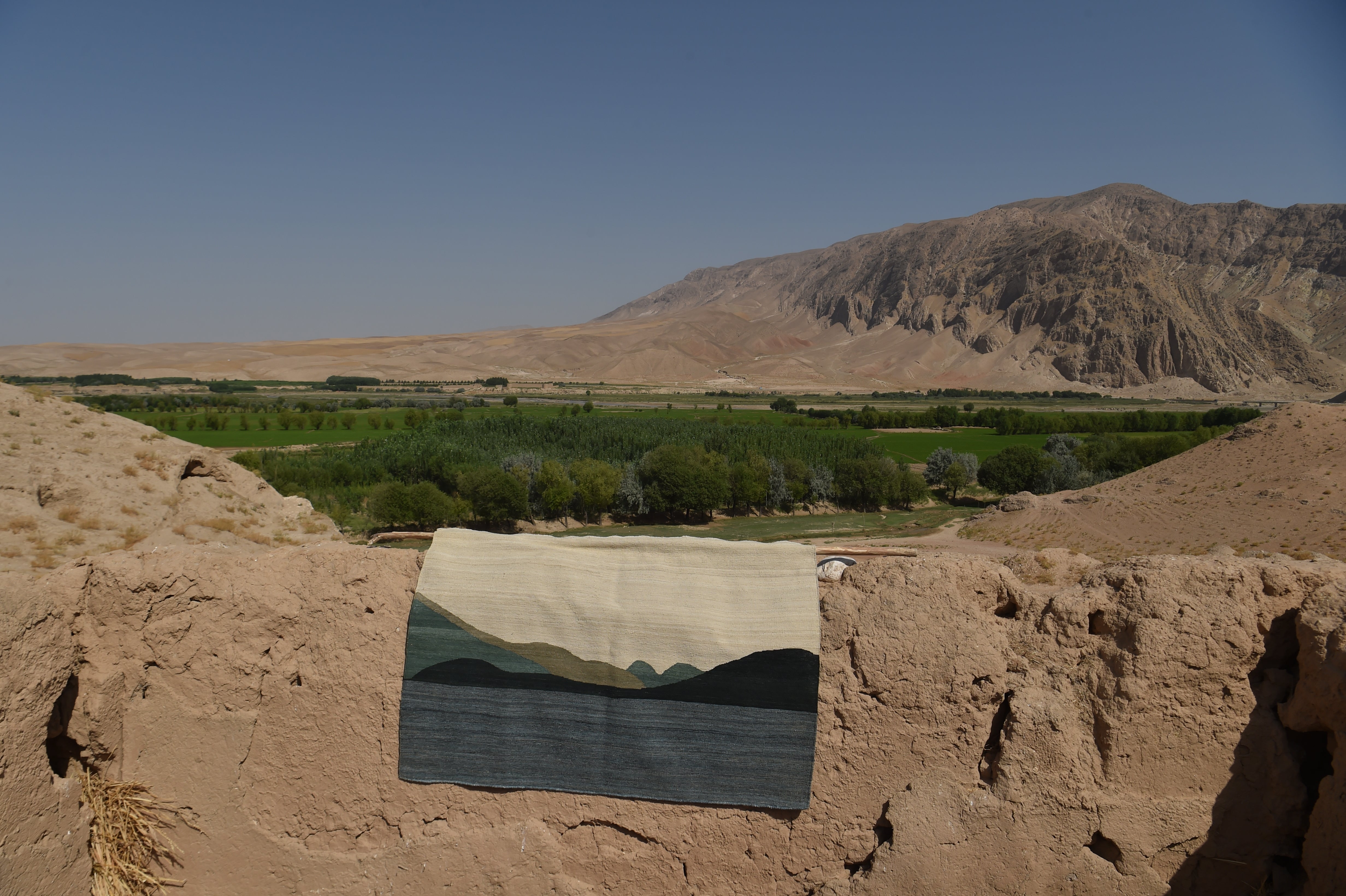 A rectangular flatweave rug featuring a landscape design with a cream top, dark blue water, and green hills, draped over a rustic earthen wall with a desert valley and mountains in the background.