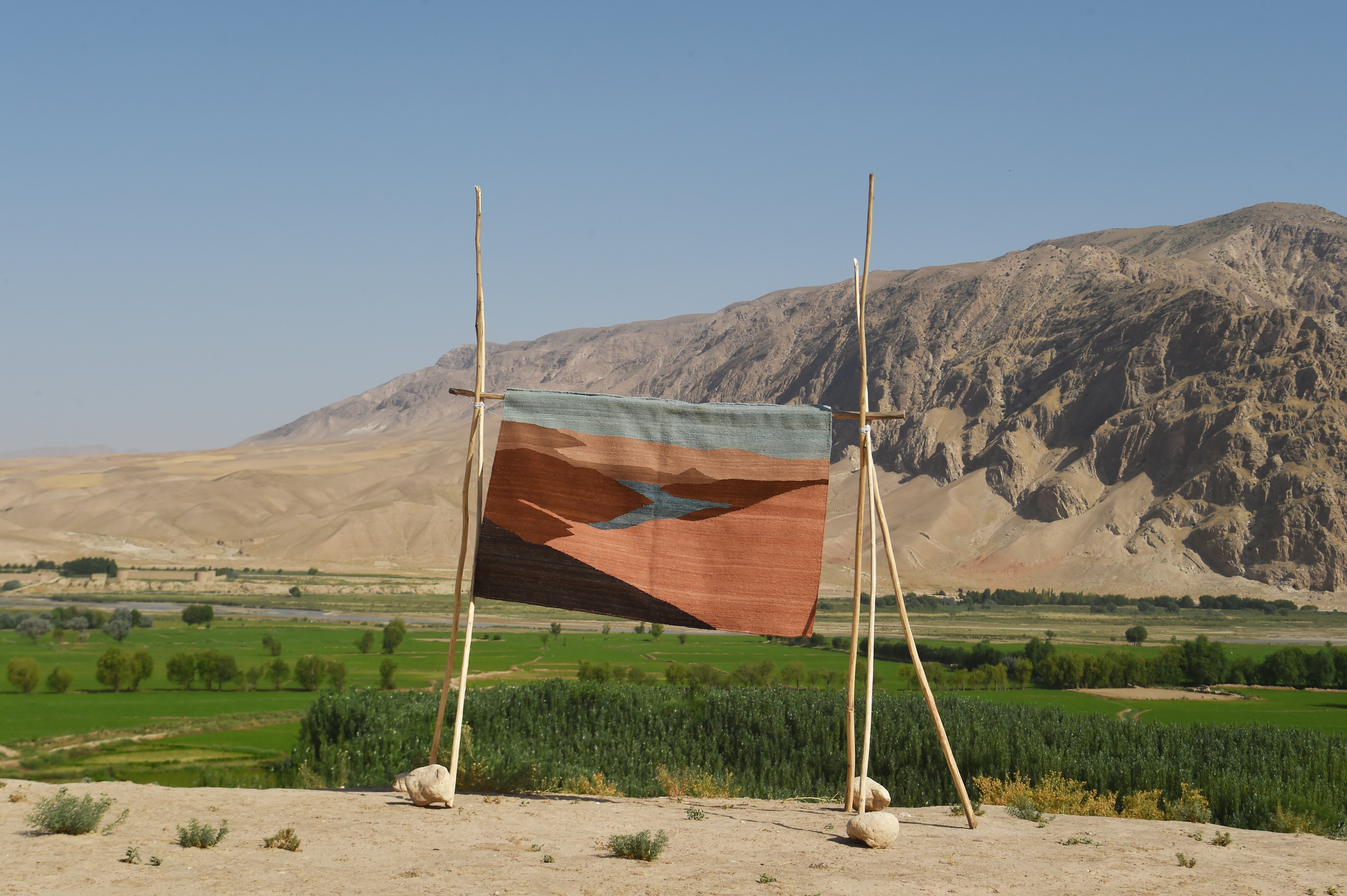 A square woven flatweave rug with a geometric pattern in terracotta, brown, and light blue, displayed on a rustic wooden tripod stand in an outdoor desert landscape with mountains and green fields in the background.
