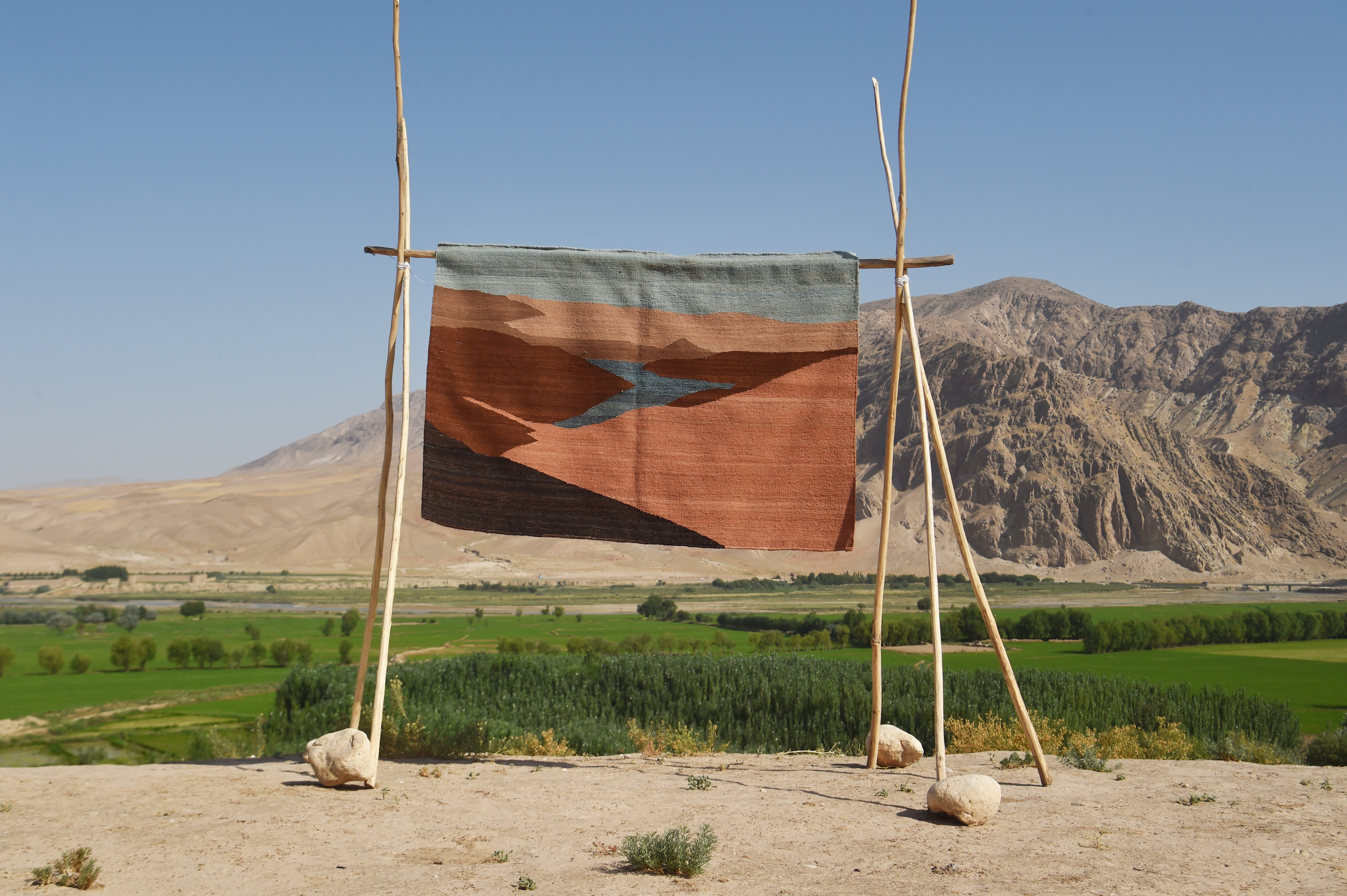 A rectangular woven flatweave rug with a geometric pattern in terracotta, brown, and slate blue hangs from a simple wooden frame outdoors, with green fields and rocky mountains in the background.