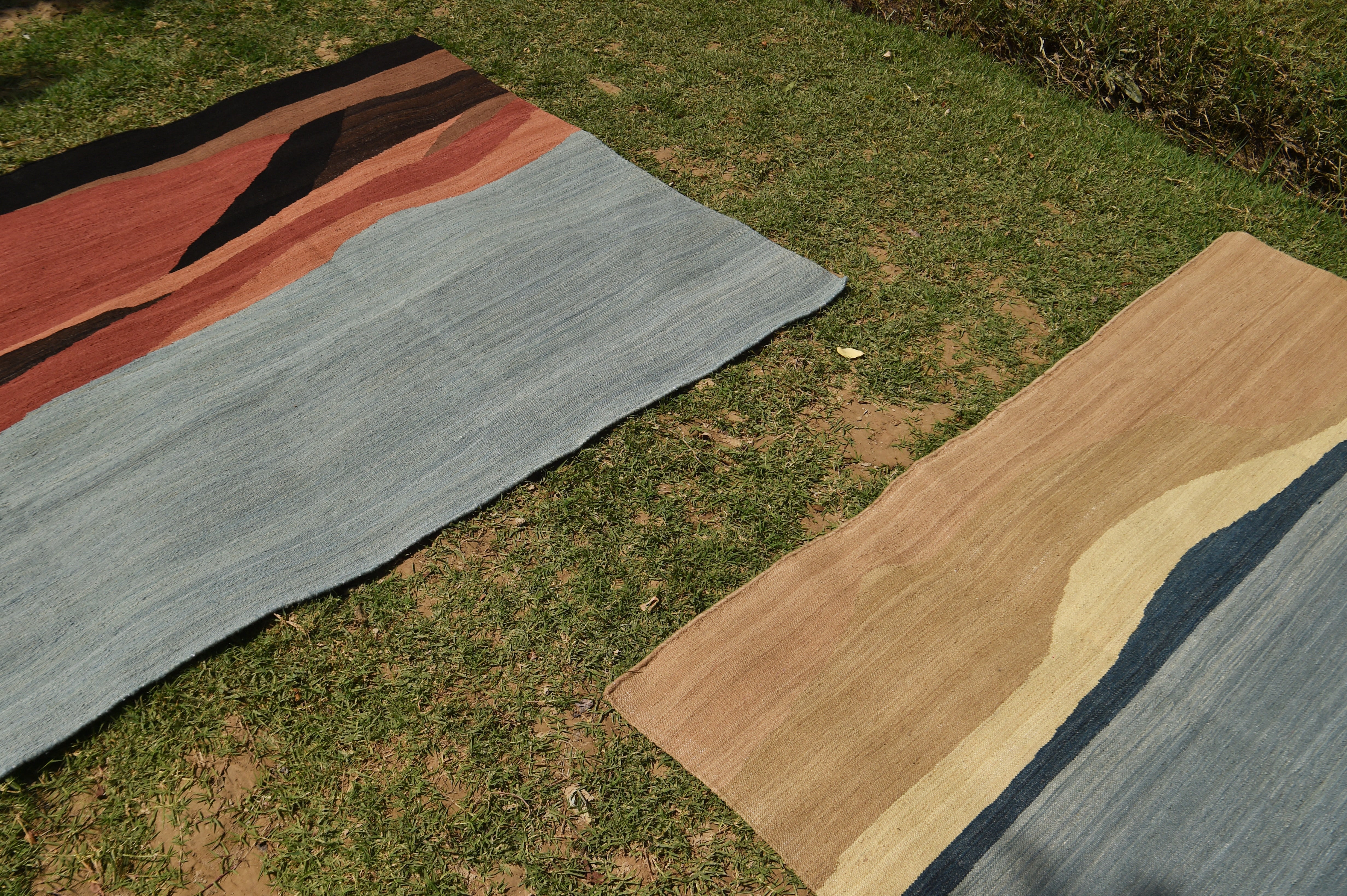 Two abstract landscape-style area rugs laid out on green grass outdoors. One rug features a large light blue section with black, rust orange, and dark brown wavy bands; the other visible rug shows tan, cream, and dark blue sections.