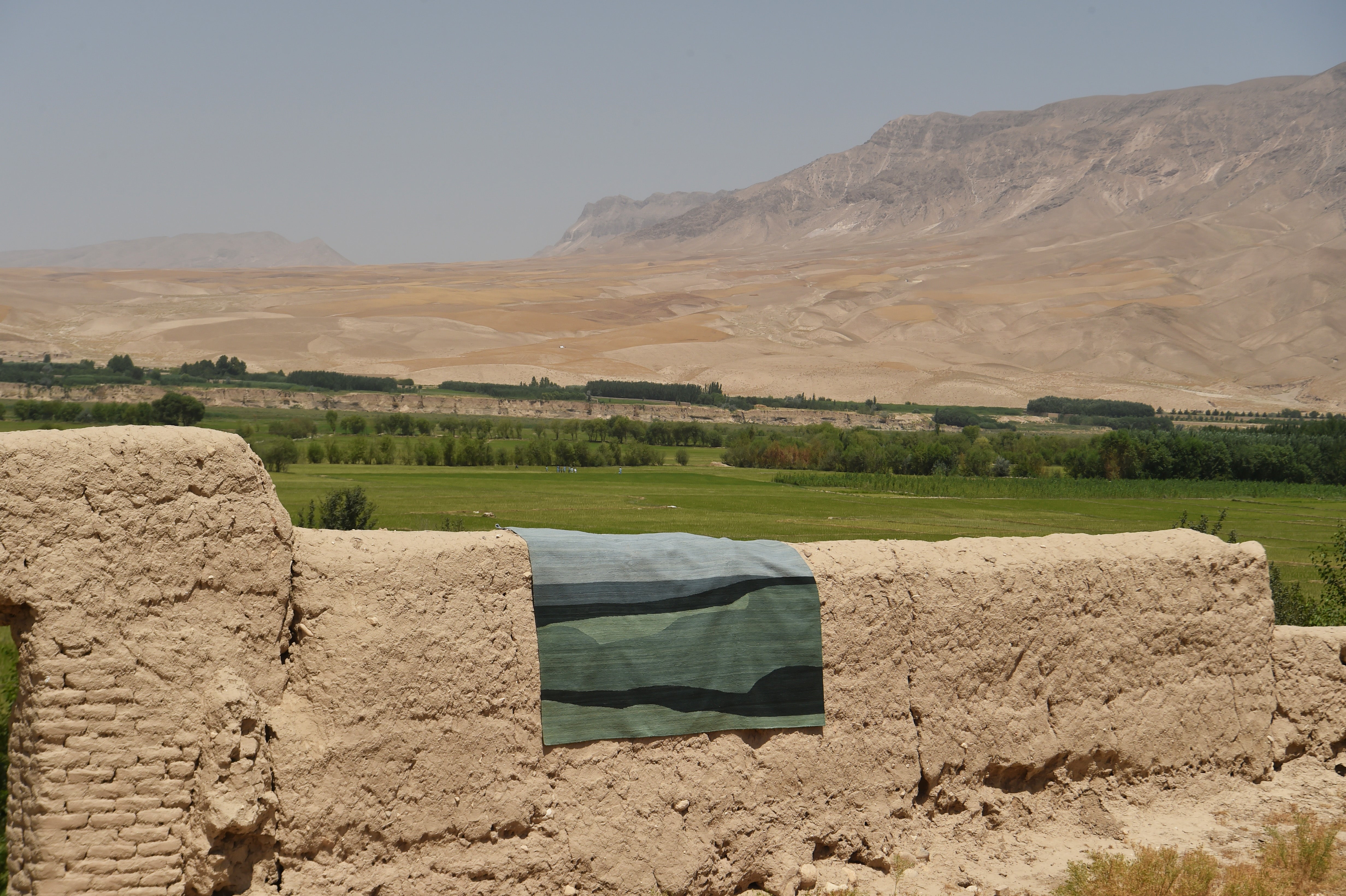 A rectangular flatweave rug with a blue and green abstract pattern draped over a rustic beige mud-brick wall, with a green valley and arid mountains in the background.