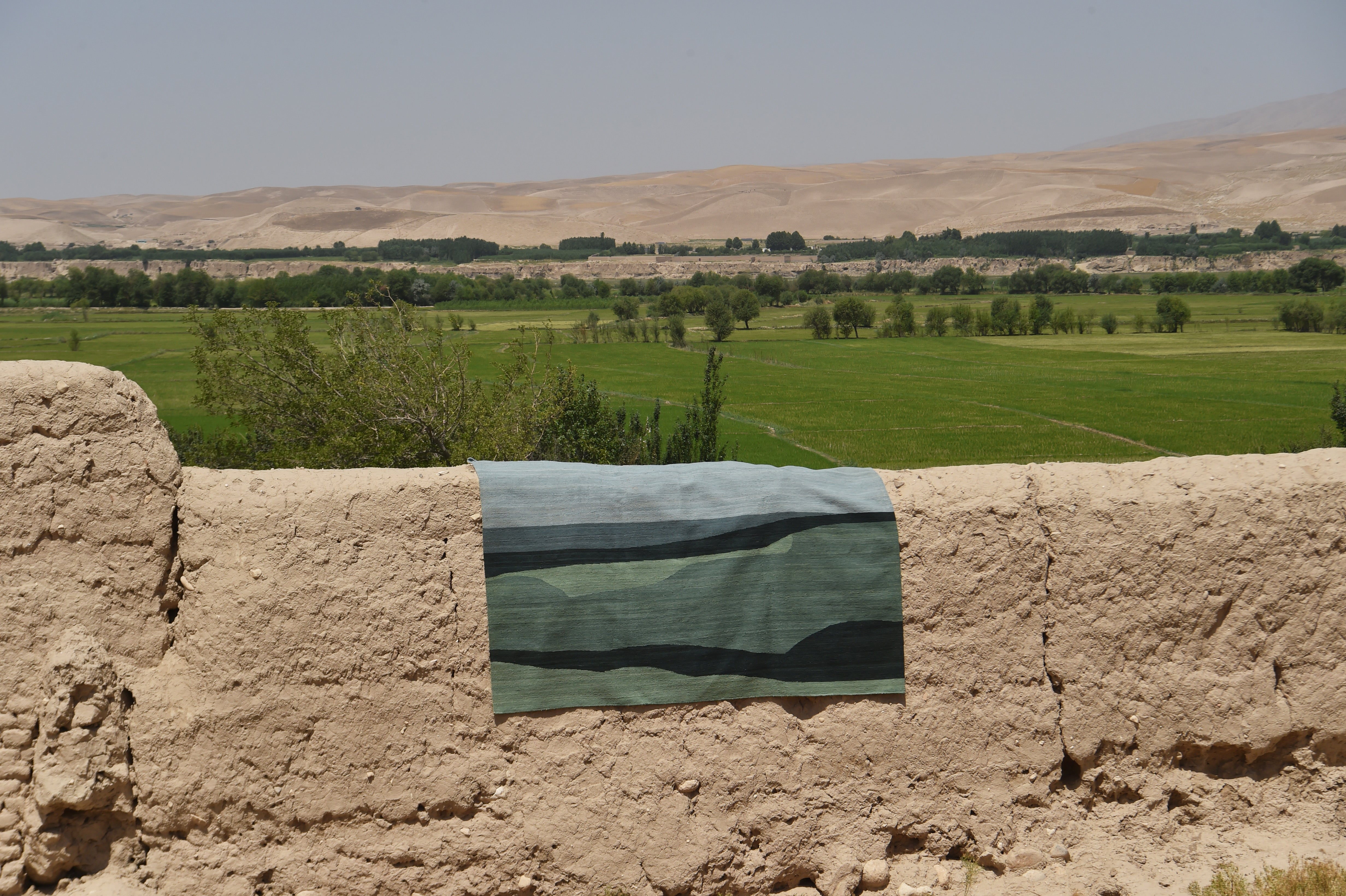 A rectangular flatweave rug with a blue and green abstract pattern draped over a rustic beige mud-brick wall, with a green valley and arid mountains in the background.