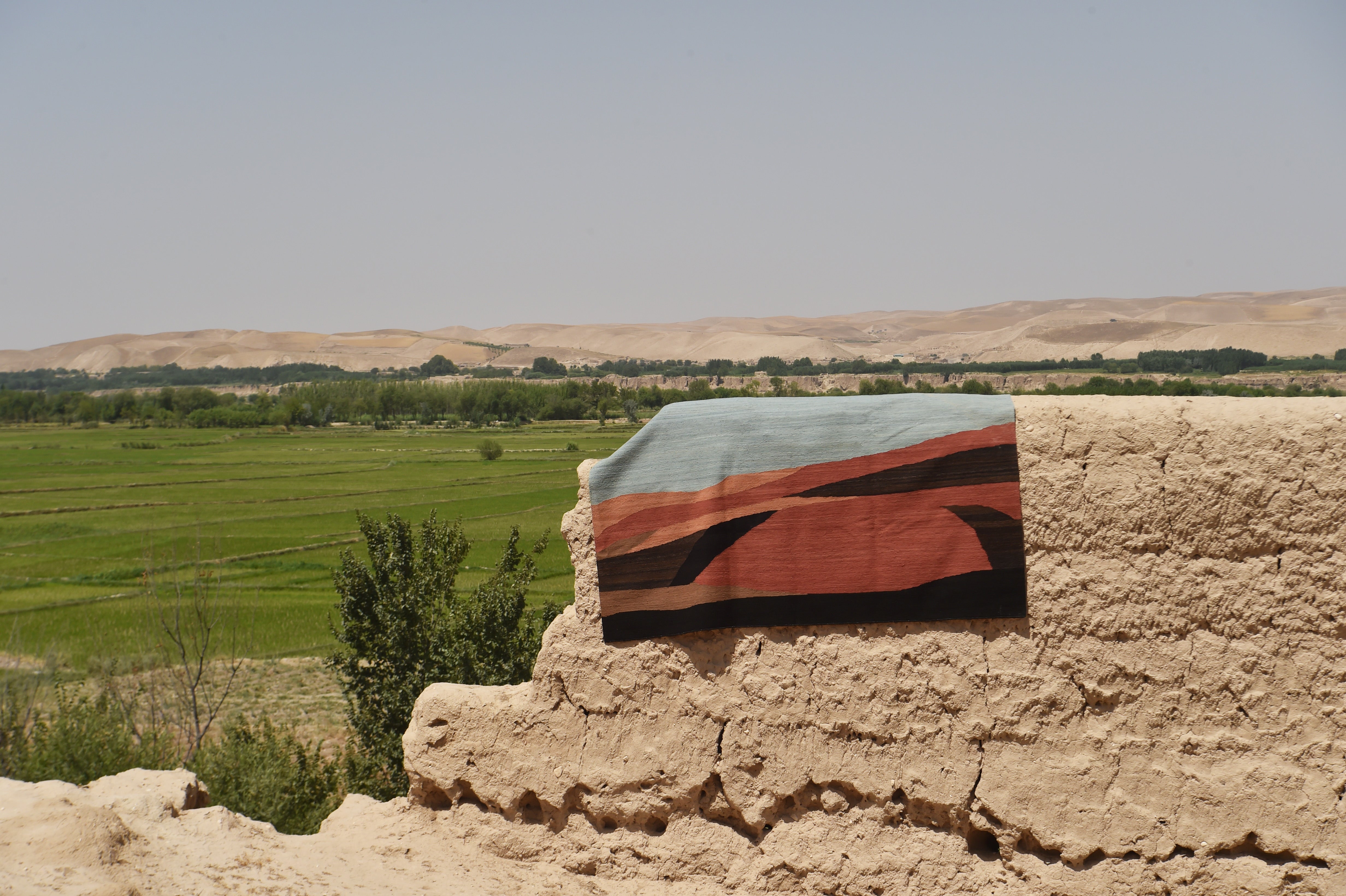 A woven  flatweave rug with a geometric pattern in light blue, rust orange, black, and beige, draped over a rustic adobe wall with a green field and desert hills in the background.