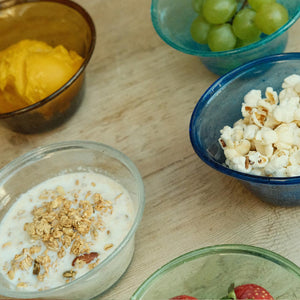 Assorted snacks including yogurt with granola, popcorn, and fruit in colourful hand-blown glass bowls