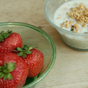 Green handblown bowl of strawberries next to a bowl of cereal with milk on a wooden surface
