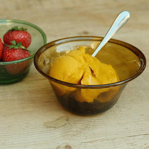 Gold hand-blown glass bowl with mango ice cream and a spoon, next to a green bowl of strawberries on a wooden surface.
