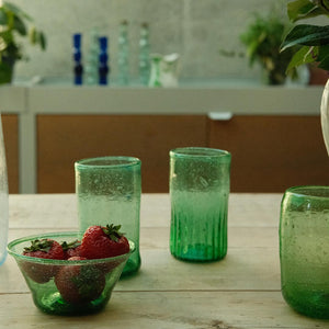 Green glass tumblers and a bowl of strawberries on a wooden table with a blurred background.