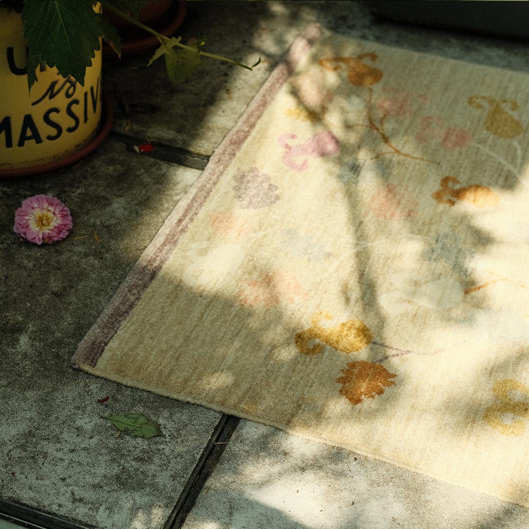 Floral-patterned rug on a concrete floor with a plant and pink flower in the background.