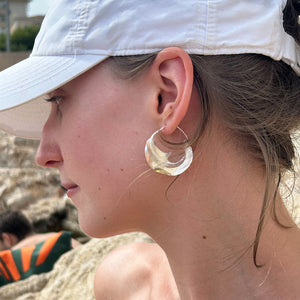 Close-up of a person wearing a white cap and large silver Malain twist earrings on a beach.
