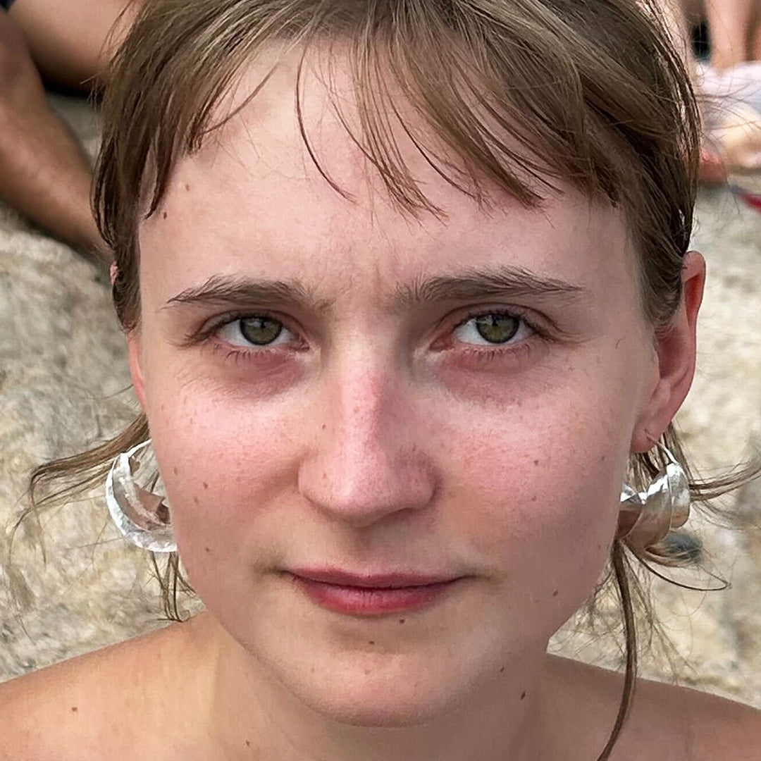 Close-up of a person wearing large silver Malain twist earrings on a beach.
