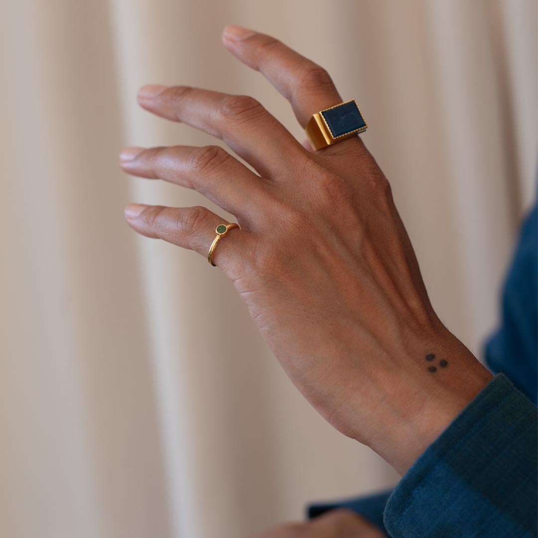 Hand wearing two gold rings with gemstones against a neutral background