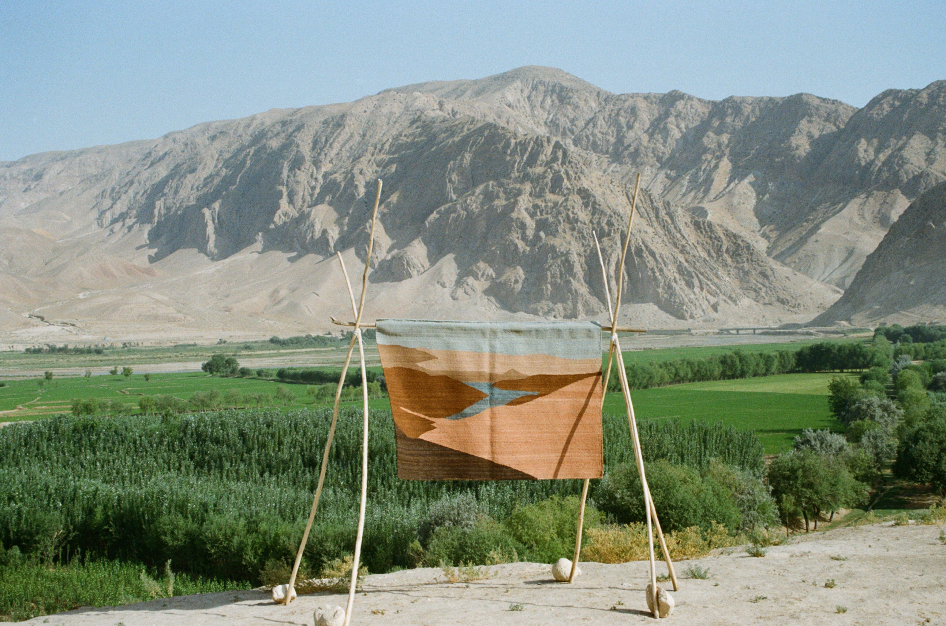 A woven textile art piece rug featuring geometric brown and beige shapes with a blue river motif, displayed on a wooden tripod frame against a scenic background of green fields and mountains.