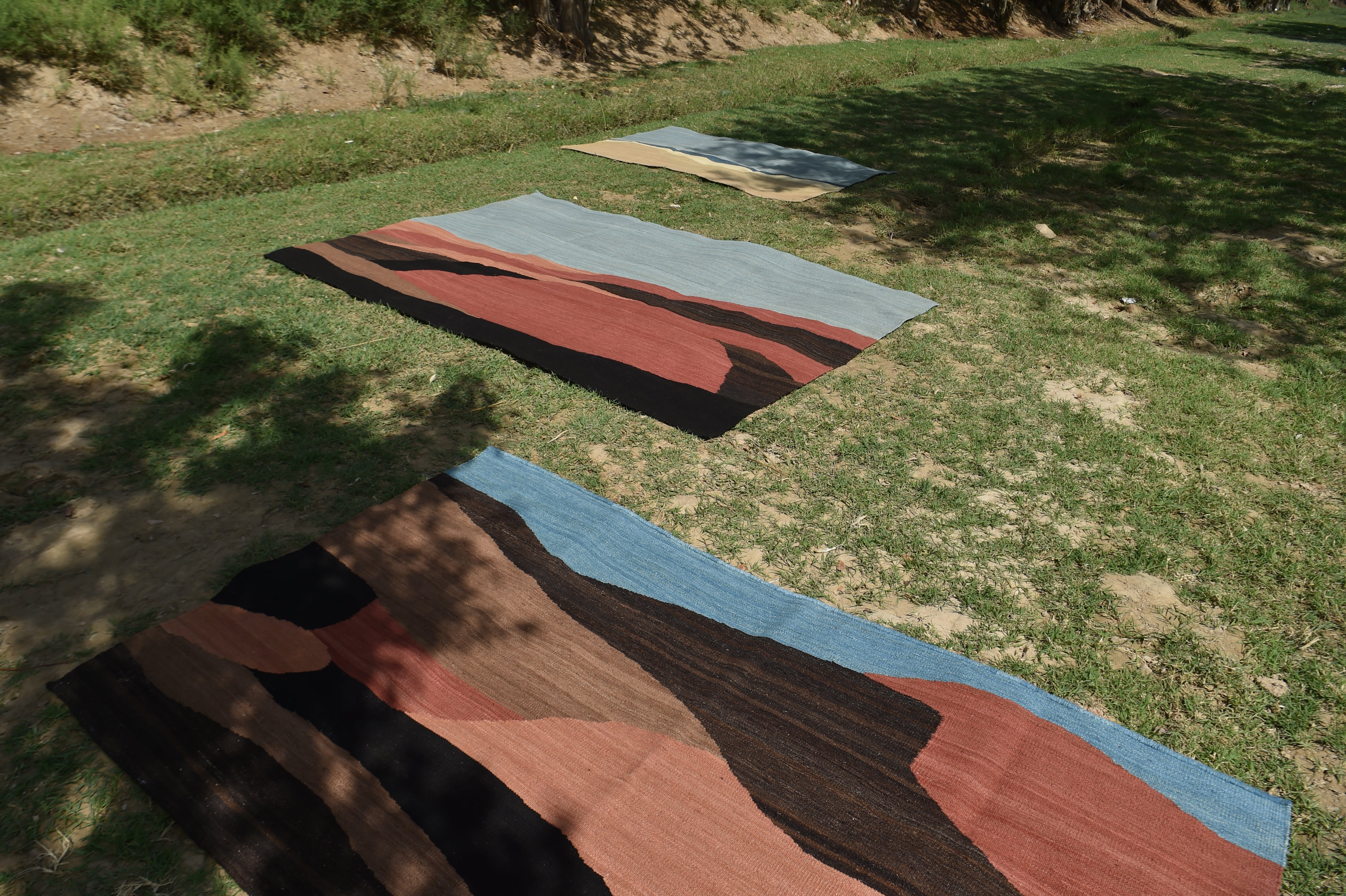 Three abstract landscape design area rugs laid out on green grass. Designs comprise colour blocks of terracotta, brown, sand and blue.