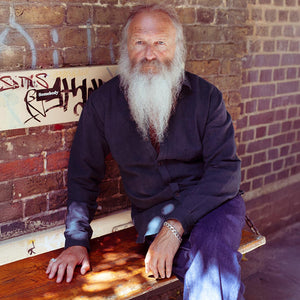 Man with a long white beard sitting on a wooden bench against a brick wall wearing a dark green cotton shirt