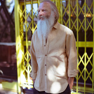 Man with a long white beard wearing a ochre cotton shirt standing in front of a yellow lattice structure