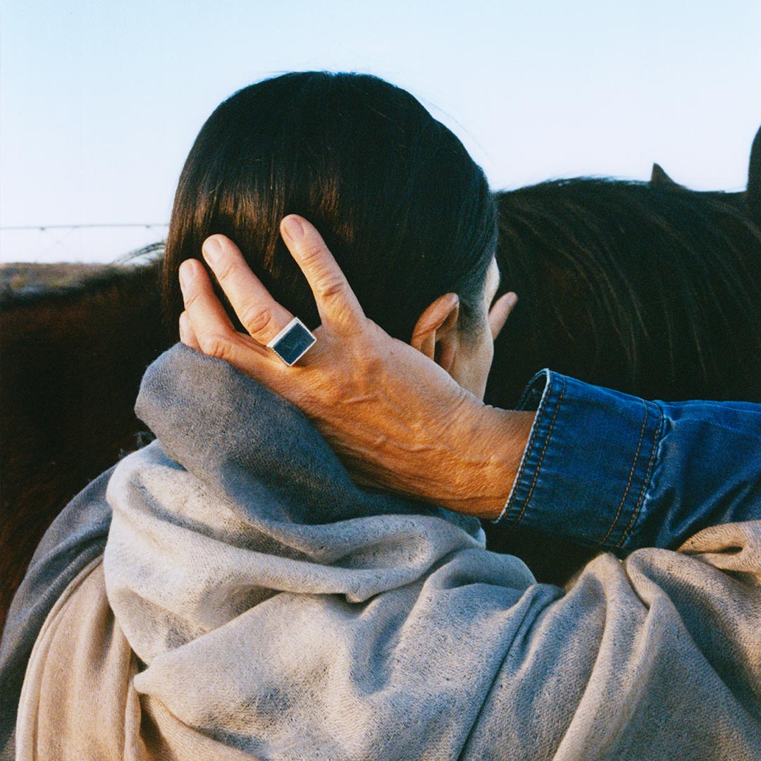 Model wearing a blue agate ring with a silhouette of a galloping horse etched onto the stone, set in a sterling silver band, next to a horse.